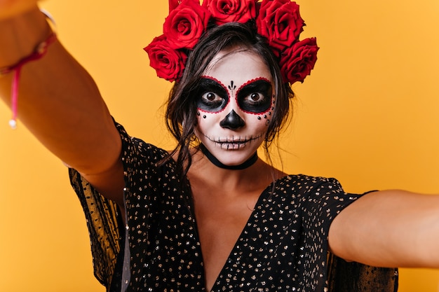 Girl with eyes wide open in surprise makes selfies. snapshot of brunette with skull mask in orange wall