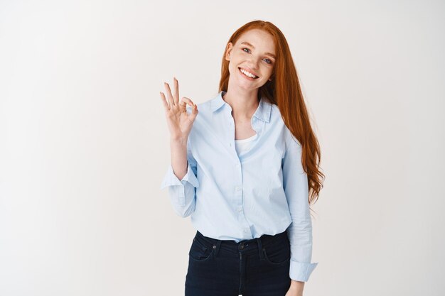 Girl with ginger hair showing OK sign and smiling, recommending your promotion product, standing satisfied against white wall