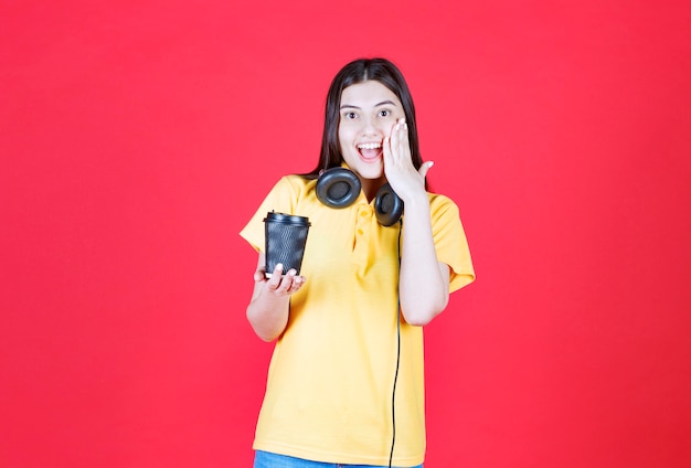 Girl with headphones holding a black disposable cup of drink, covering mouth and looks surprized.