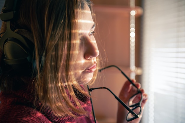 A girl with headphones behind a laptop in the sunlight through the blinds