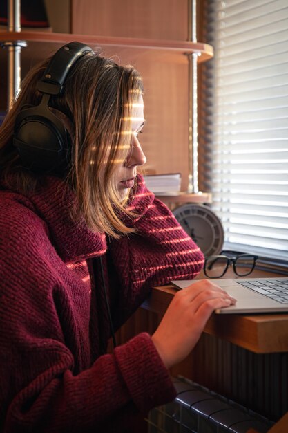 A girl with headphones behind a laptop in the sunlight through the blinds