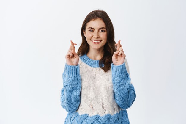 Girl with high hopes making wish, cross fingers good luck and smiling, praying or hoping for positive result, standing against white wall