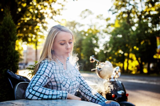 Девушка с длинными светлыми волосами в футболке сидит за столом снаружи и курит электронную сигарету.