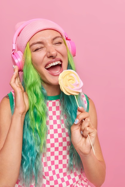 girl with long dyed hair holds delicious candy on stick listens favorite song in headphones wears casual dress and hat isolated on pink 