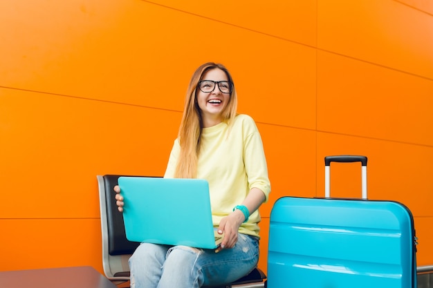Girl with long hair in yellow sweater is sitting on orange background. She has blue suitcase and laptop. She is smiling happy.