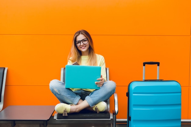 Girl with long hair in yellow sweater is sitting on orange background. She has blue suitcase and laptop. She is smiling happy.