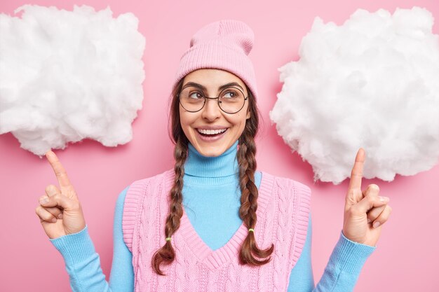 흰 구름에 위의 땋은 머리를 가진 소녀는 뭔가 모자 터틀넥 라운드 안경과 분홍색에 고립 된 조끼를 착용하는 것을 보여줍니다