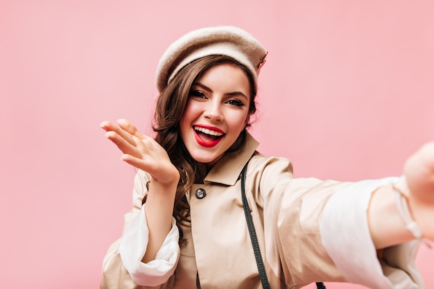 Girl with red lips dressed in beige trench coat and hat blows kiss and takes selfie on pink background.