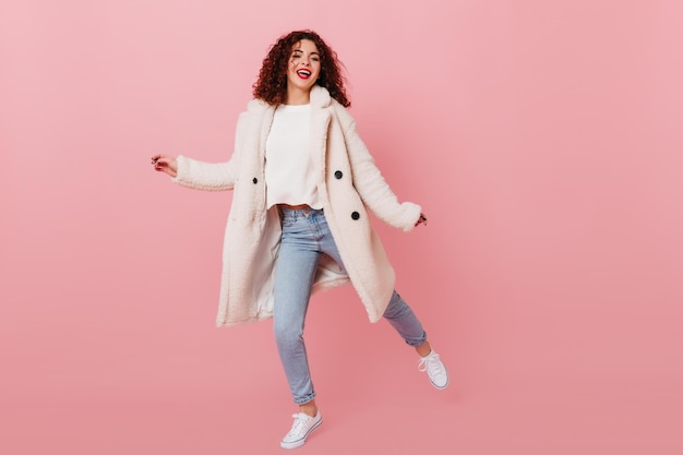 Girl with red lips is dancing on pink background Woman with dark curly hair dressed in warm fur coat and denim skinny pants smiling