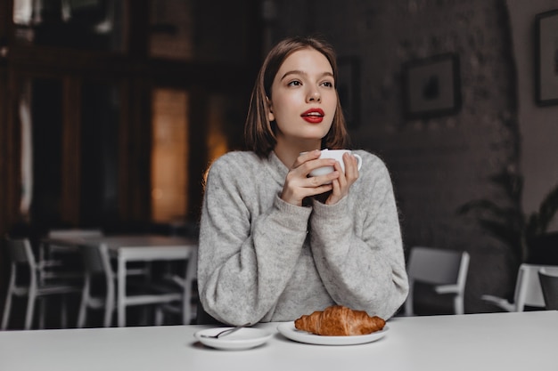 Girl with short hair and red lipstick dressed in warm sweater enjoying tea with croissant in cozy cafe.