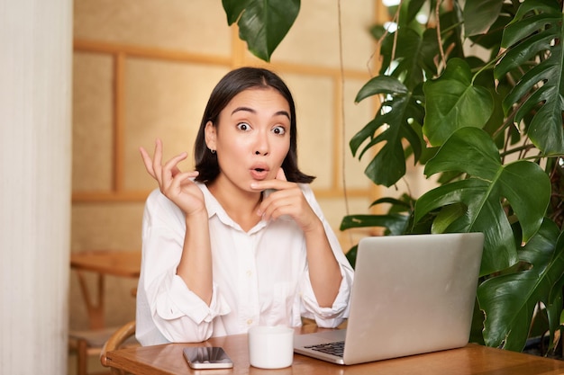 Foto gratuita ragazza con il viso sorpreso che lancia un'idea che punta a sinistra seduta con il computer portatile in un caffè e beve caffè