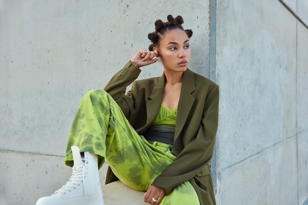 girl with trendy hairstyle dressed in green clothes looks away poses against urban grey wall considers something