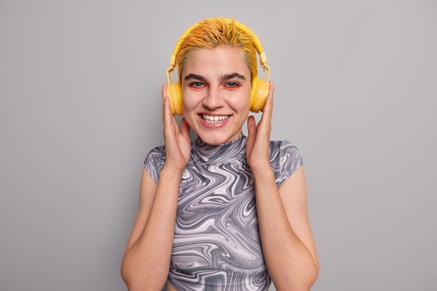 girl with trendy yellow hairstyle bright makeup listens music in wireless headphones has cheerful mood enjoys favorite rock song dressed casually on grey