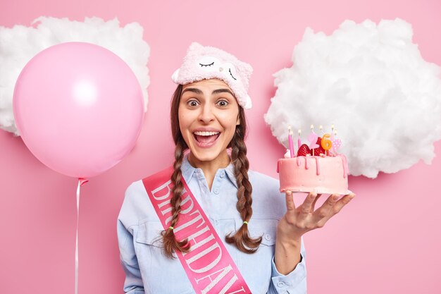 girl with two pigtails happy to accept congratulations holds delicious strawberry cake inflated helium balloon isolated on pink 