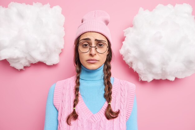 girl with two pigtails has gloomy expression sulking and frowning feels disappointed or upset wears round spectacles pink hat turtleneck and vest poses indoor