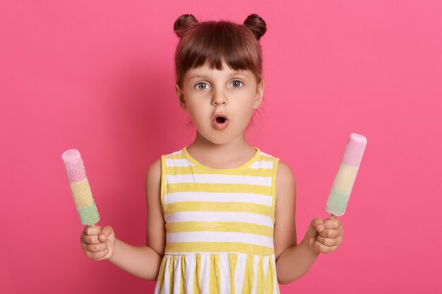 Girl with water ice creams in both hands wearing striped dress, having two knots, posing with astonished facial expression against pink wall.
