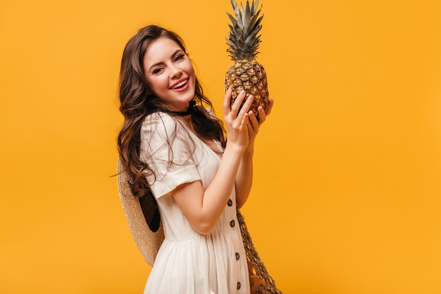 Girl with wavy dark hair is smiling and holding pineapple on orange background.