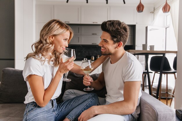 Girl with wavy hair looking at boyfriend while drinking wine. Indoor portrait of romantic couple enjoying date.