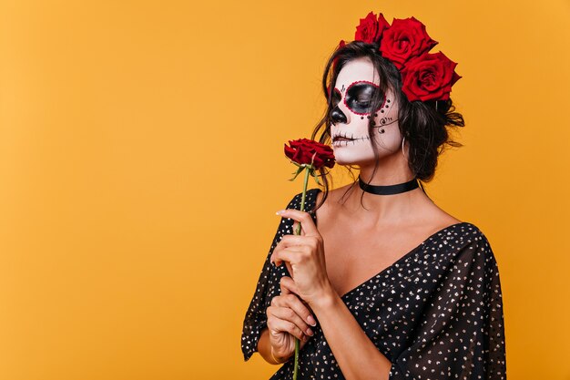 Girl with zombie mask for Halloween smells fragrant rose. Model in black dress on orange wall
