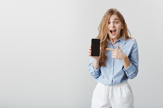 Girl won smartphone in lottery. Portrait of amazed charming young female in blue blouse showing smartphone and pointing at device with index finger, dropping jaw, yelling from excitement and surprise