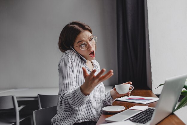 眼鏡をかけた少女労働者は、ラップトップと一杯のコーヒーを持ってオフィスに座って、感情的かつ憤慨して電話で話します。