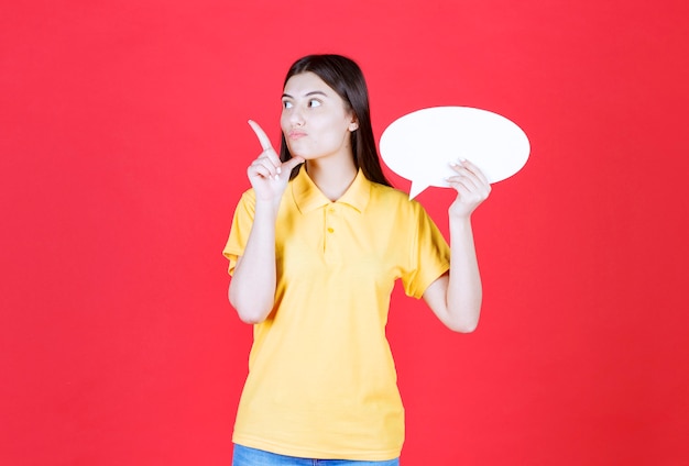 Girl in yellow dresscode holding an ovale info board and looks thoughtful and hesitating.