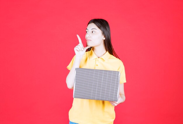 Free photo girl in yellow shirt holding silver gift box and looks confused and thoughtful