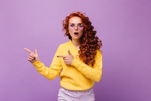 Girl in yellow sweater and lilac glasses looks surprised at camera on isolated wall