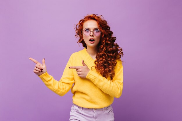 Girl in yellow sweater and lilac glasses looks surprised at camera on isolated wall