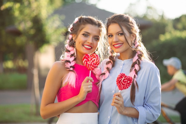 Girlfriends with colorful braids holding candy heart on stick.