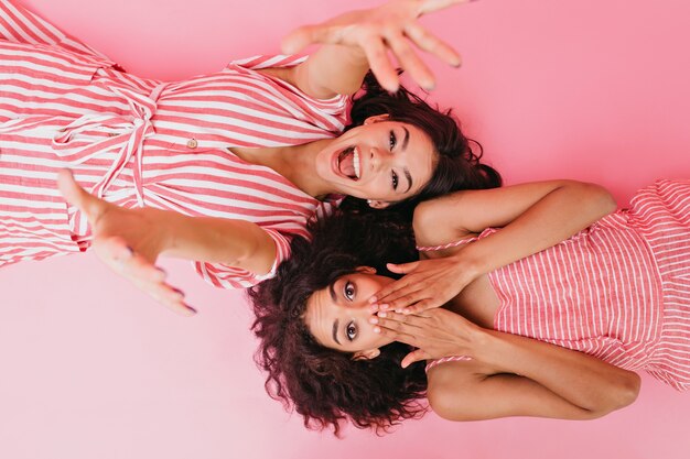 Girlfriends with dark curly hair are having fun at their pajama party. Laughing girl stretches towards while her cousin covers her face in surprise.