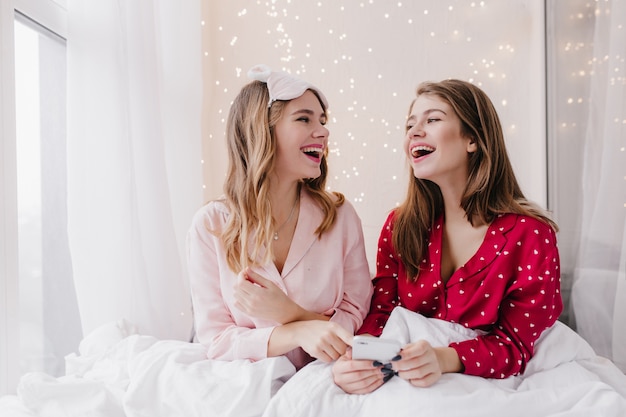 Девушки в ночных костюмах смотрят друг на друга и улыбаются. Фото в помещении смеющихся кавказских дам, сидящих в постели со смартфоном.