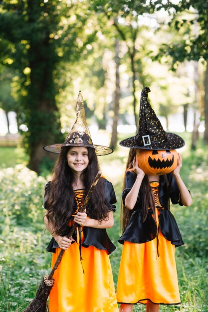 Girls in sorceress costumes holding broomstick and closing face by pumpkin