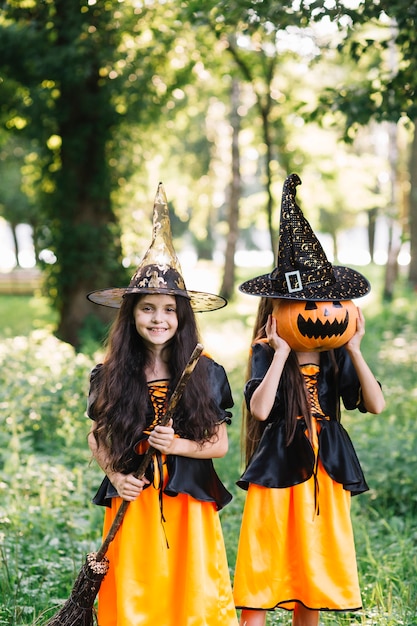 Girls in sorceress costumes holding broomstick and closing face by pumpkin