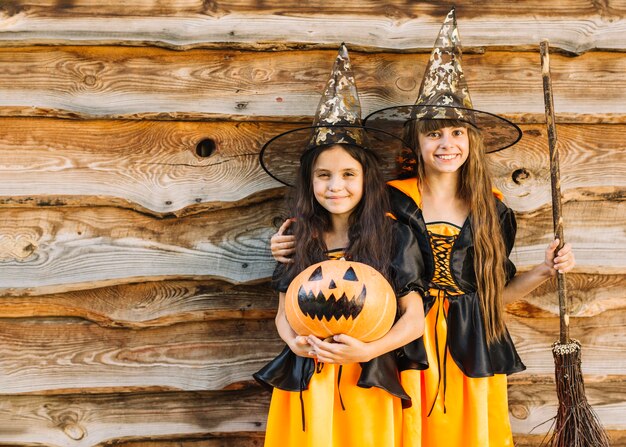 Girls in witch costumes hugging, smiling, holding broomstick and pumpkin