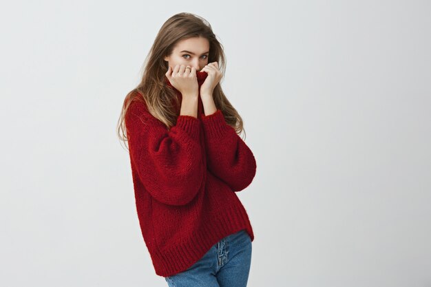 Give me cuddle to warm up. Portrait of cute good-looking european woman hiding face in collar of sweater glancing  while standing . Girl freezes because of broken heater