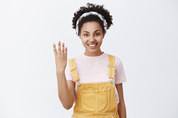 Give me four. Portrait of friendly-looking caring and cute elder sister with dark sking in trendy yellow overalls and headband over hair showing fourth number with fingers and smiling with kind smile