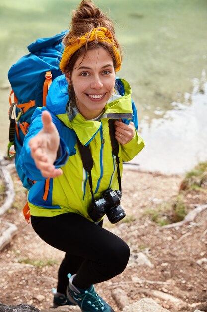 手を貸して！満足している女性の行楽客の垂直ショットは、丘を登ろうとし、ターコイズブルーの湖の近くでポーズをとって、カメラで手を伸ばします