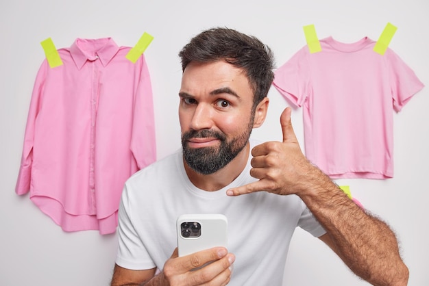 Give me your telephone number Serious bearded man makes call me gesture uses mobile phone dressed in casual t shirt poses against white background with hanging clothes behind Body language