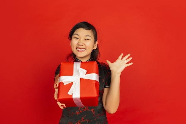 Дарить подарочную коробку. Счастливый китайский Новый год 2020. Портрет азиатской молодой девушки, изолированные на красном фоне. Женская модель в традиционной одежде выглядит счастливой. Праздник, праздник, эмоции. Copyspace.
