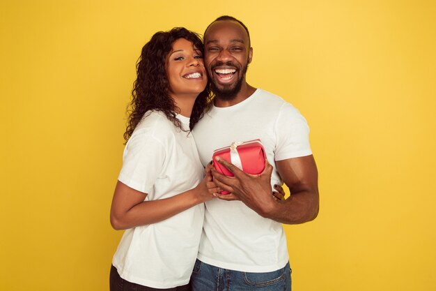 Giving surprise. Valentine's day celebration, happy african-american couple isolated on yellow studio background. Concept of human emotions, facial expression, love, relations, romantic holidays.