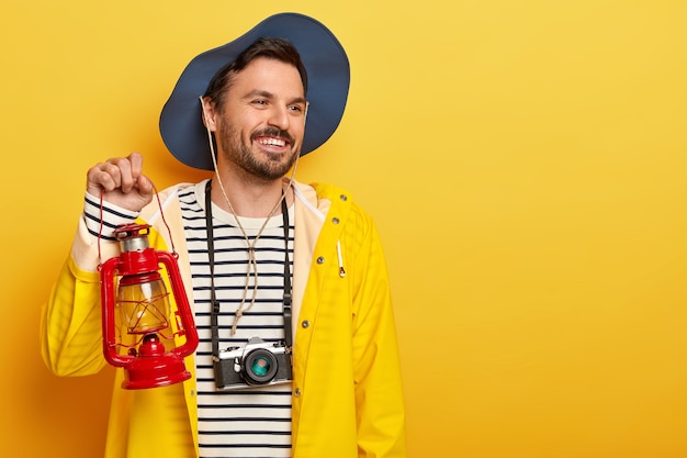 Glad active male holds gas lamp, has walk in forest, wears retro camera on neck, looks with smile aside, wears hat and raincoat, poses over yellow wall