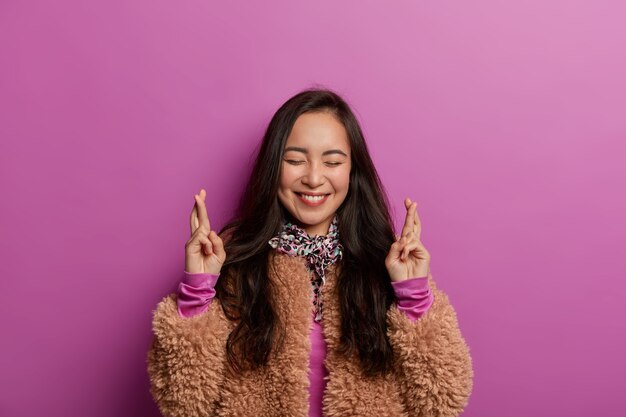 Glad adorable Asian woman waits for special moment, keeps fingers crossed, prays for better, smiles toothily with closed eyes