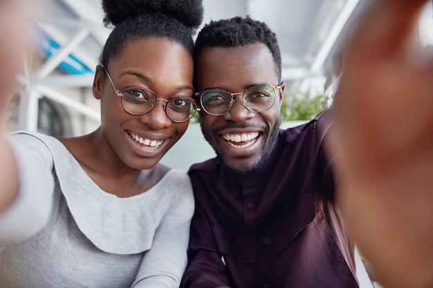 Glad African friends enjoy resting together, have positive expressions, make photo, stretch hand as take selfie, share pictures in social networks.