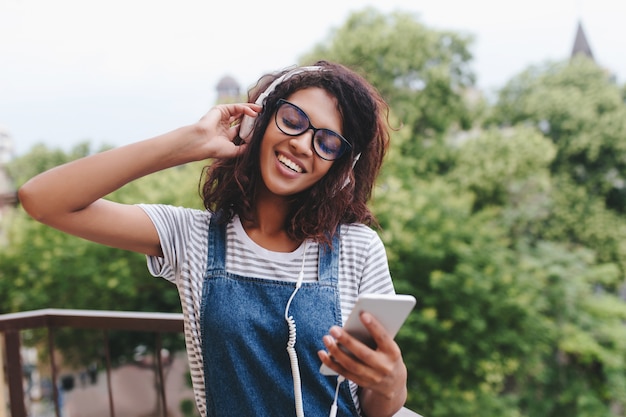 Glad african young woman in striped shirt standing on tree and listening song with smile and eyes closed