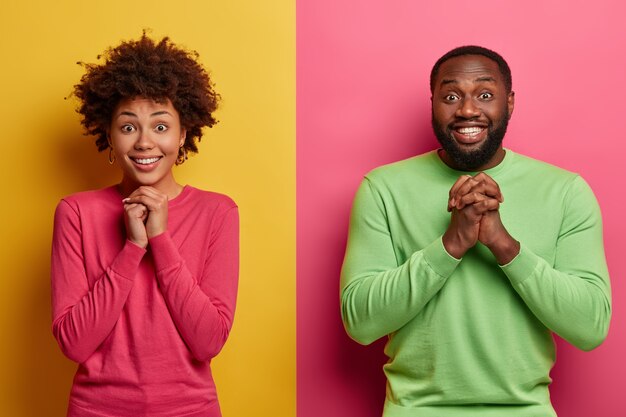 Glad Afro American girlfriend and boyfriend keep hands together in praying gesture, anticipate important results, stand next to each other against two colored wall, smile broadly, feel happy