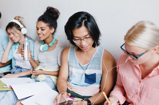 Glad asian student typing message on smartphone while his charming blonde friend writing lecture. Indoor group portrait of college mates with boy and girl in earphones.