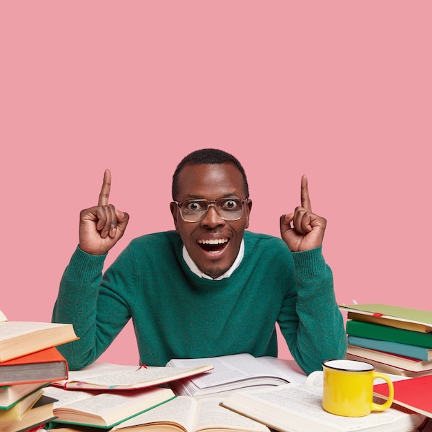 Glad astonished black male in green jumper points with both index fingers, has toothy smile, surrounded with many books