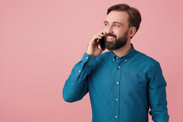 Felice attraente uomo barbuto, distoglie lo sguardo e sorride, indossa una camicia di jeans, parla al telefono. isolato su sfondo rosa.