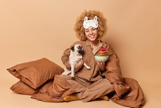 Glad beautiful woman poses with favorite pug dog and appetizing pancakes in bed start day with eating something delicious wears sleepmask and pajama isolated over beige background Morning time